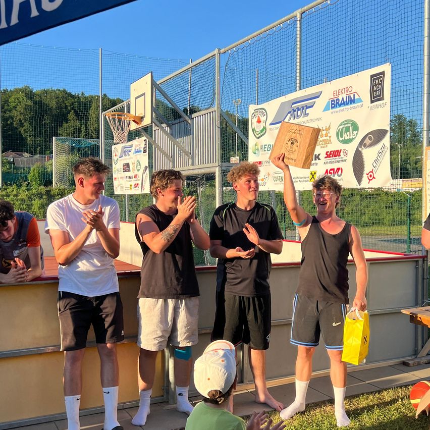 Fünf Männer stehen auf einer Plattform vor einer Menge und halten eine Trophäe und eine Tasche. Sie scheinen einen Sieg zu feiern. Dahinter sind ein Banner und ein Basketballkorb zu sehen.