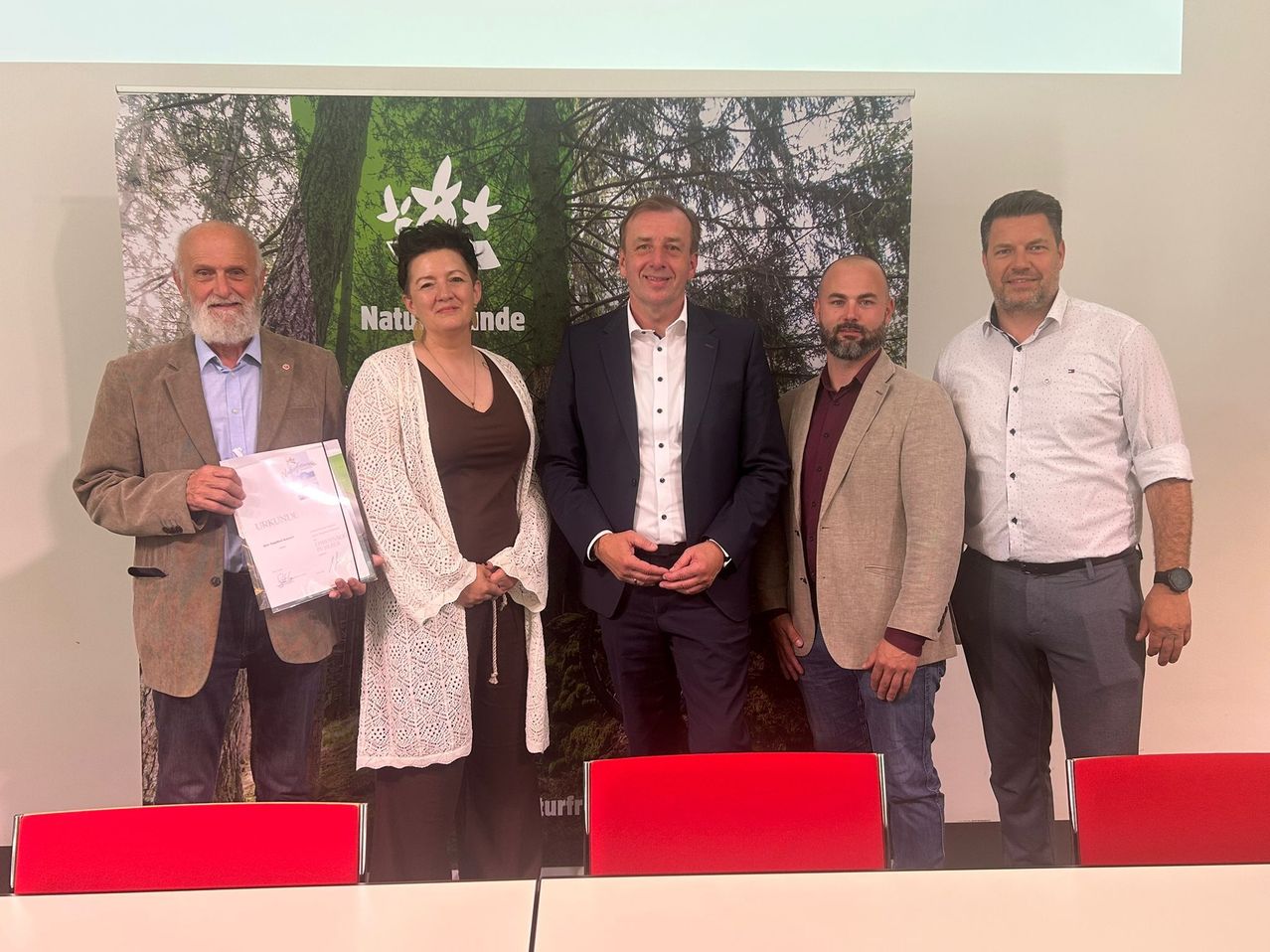 Fünf Personen stehen vor einem grünen Hintergrund und lächeln. Der Mann links hält ein Zertifikat. Hinter ihnen befindet sich ein großes Poster mit einer Waldlandschaft.