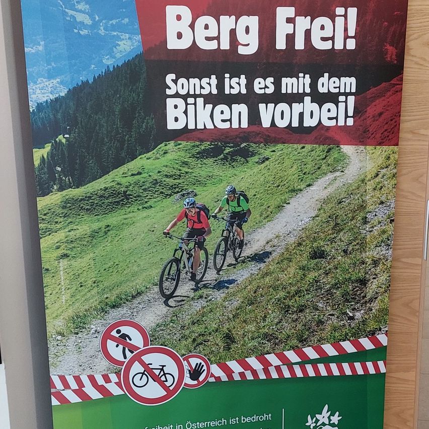 Ein Plakat zeigt zwei Radfahrer auf einem Bergpfad mit dem Slogan 'Berg Frei!' und einer Warnung vor dem Radfahren auf dem Pfad.