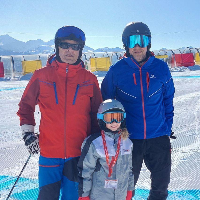 Three people stand on a snowy slope with mountains in the background. Two men wear ski jackets and helmets, one in red and one in blue. A child in a gray jacket stands between them, smiling and wearing goggles.