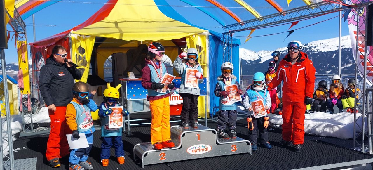 Several children stand on a podium with numbers 1, 2, and 3, holding trophies. They wear winter gear, including helmets and goggles. Behind them is a colorful tent and snowy mountains.