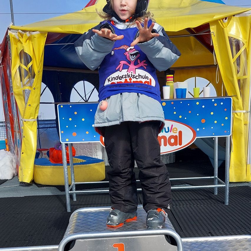 A young child stands on a podium under a colorful tent, wearing a ski school shirt and ski boots, with a table and cups behind them.