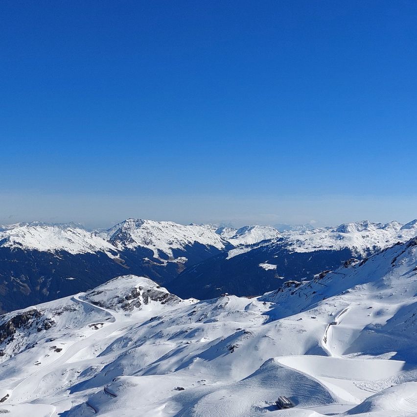 A high-altitude view of snow-covered mountains under a clear blue sky. The terrain features gentle slopes and ridges, with some tracks visible on the snow.