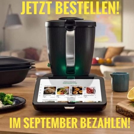 Ein Küchengerät auf einem Tisch mit einem Teller Essen, Zitronen und einem Krug. Das Gerät hat einen Bildschirm mit Rezepten. Darüber steht der Text 'JETZT BESTELLEN!' Unten ist 'IM SEPTEMBER BEZAHLEN!' geschrieben. Dahinter sind eine Couch, ein Tisch und eine Lampe zu sehen.