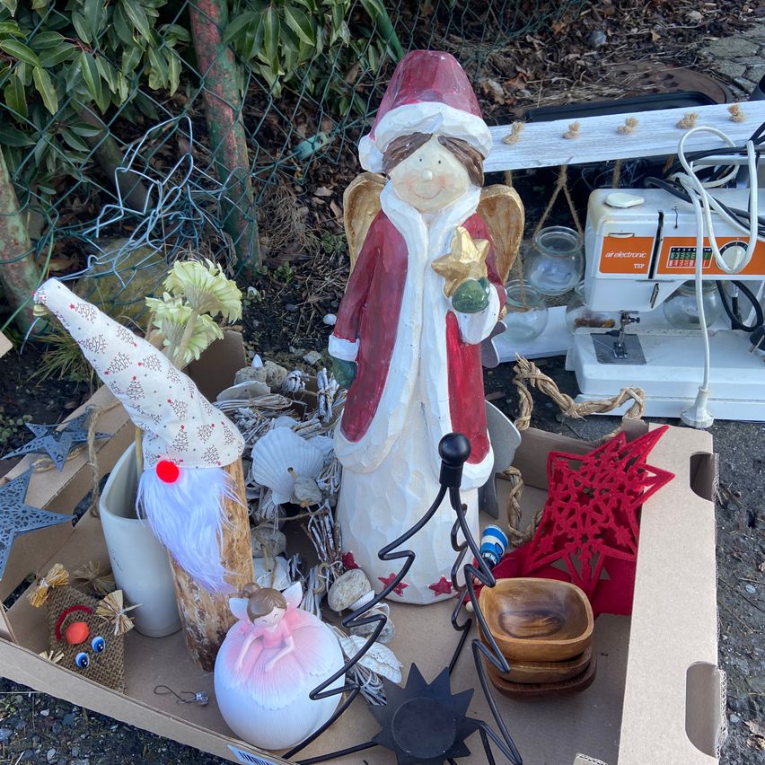 Eine Schachtel mit Weihnachtsdekorationen, darunter eine Weihnachtsmann-Figur, ein Engel, ein Elf und Ornamente, draußen an einem sonnigen Tag ausgestellt.