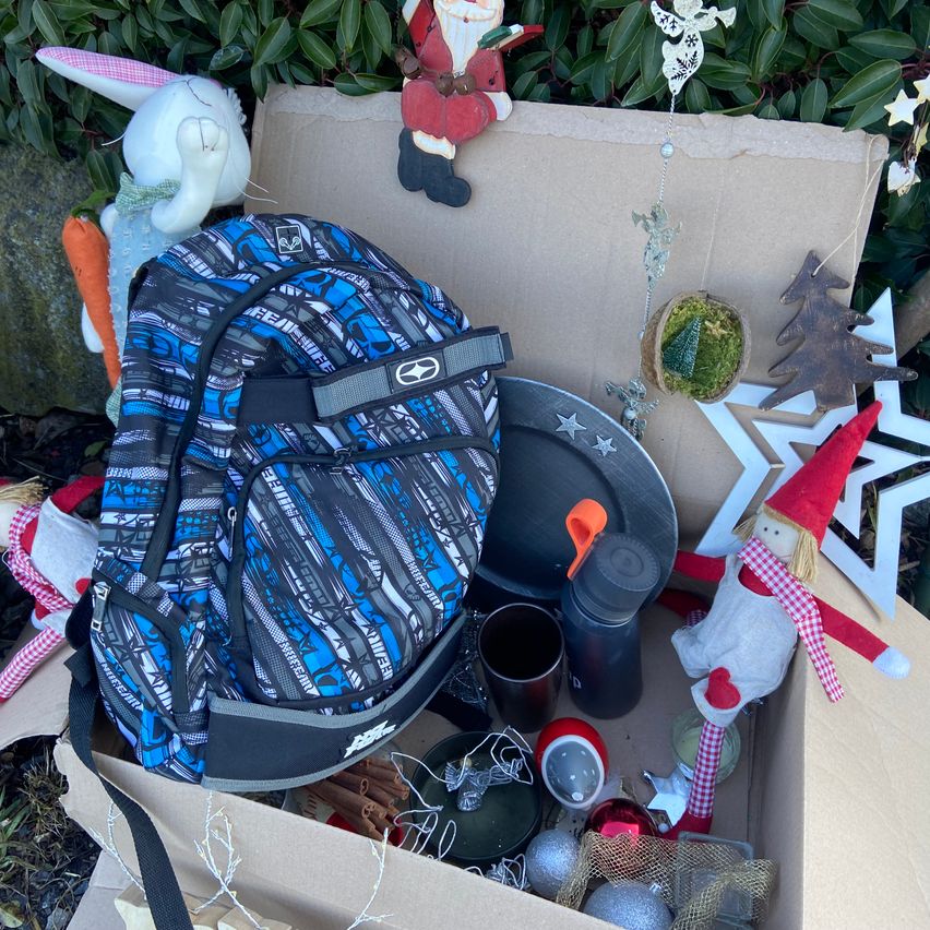 Ein Pappkarton gefüllt mit verschiedenen Gegenständen wie einem blauen Rucksack, Weihnachtsdekorationen, einer Karotte, einer Weihnachtsmann-Figur, einem Teller und einer Flasche.