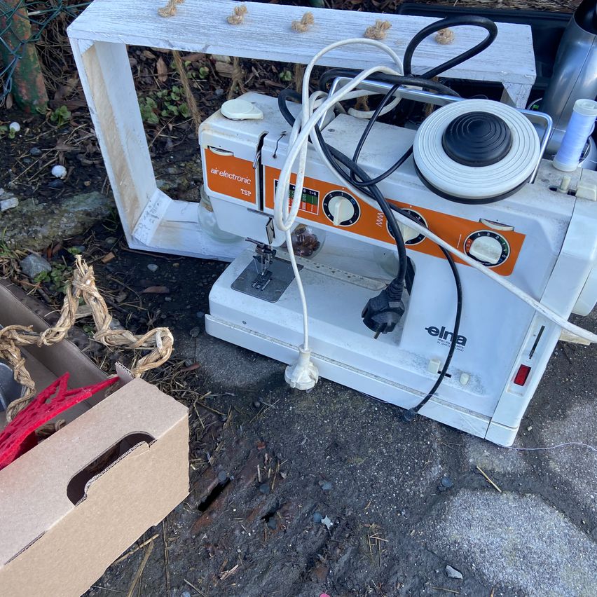 Eine alte Elna-Nähmaschine liegt neben einem Karton mit roten Objekten auf dem Boden. Die Maschine hat angeschlossene Kabel.