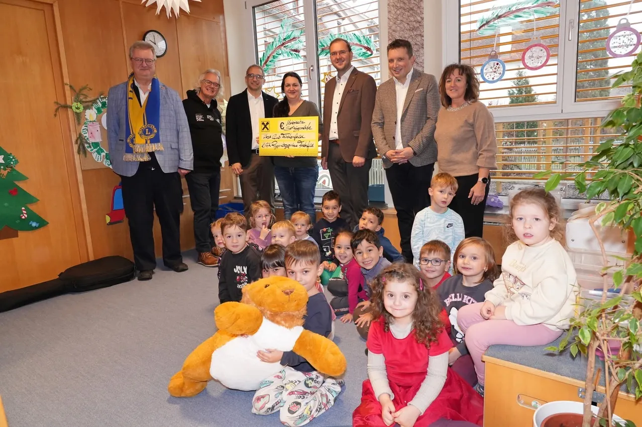 Mehrere Erwachsene und Kinder sind drinnen, einige stehen, andere sitzen auf dem Boden. Ein Mann hält ein gelbes Banner mit Text. Uhren und eine Weihnachtsdekoration hängen an der Wand.
