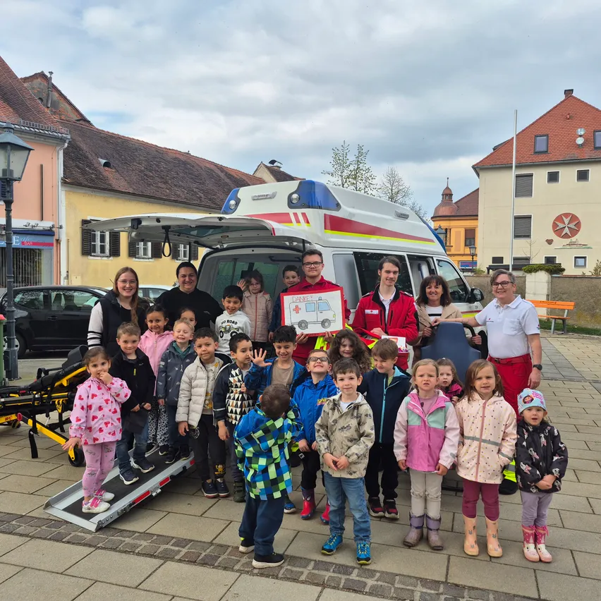 Eine Gruppe von Kindern und Erwachsenen steht um einen Krankenwagen mit offenem Heck herum und posiert für ein Foto. Der Ort scheint ein Stadtplatz zu sein.