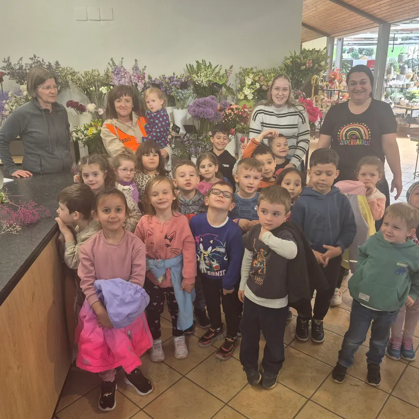 Eine Gruppe von Kindern und Erwachsenen steht vor einem Tresen mit Blumen. Einige lächeln, während andere neugierig schauen. Die Erwachsenen tragen Jacken und halten ein Baby.