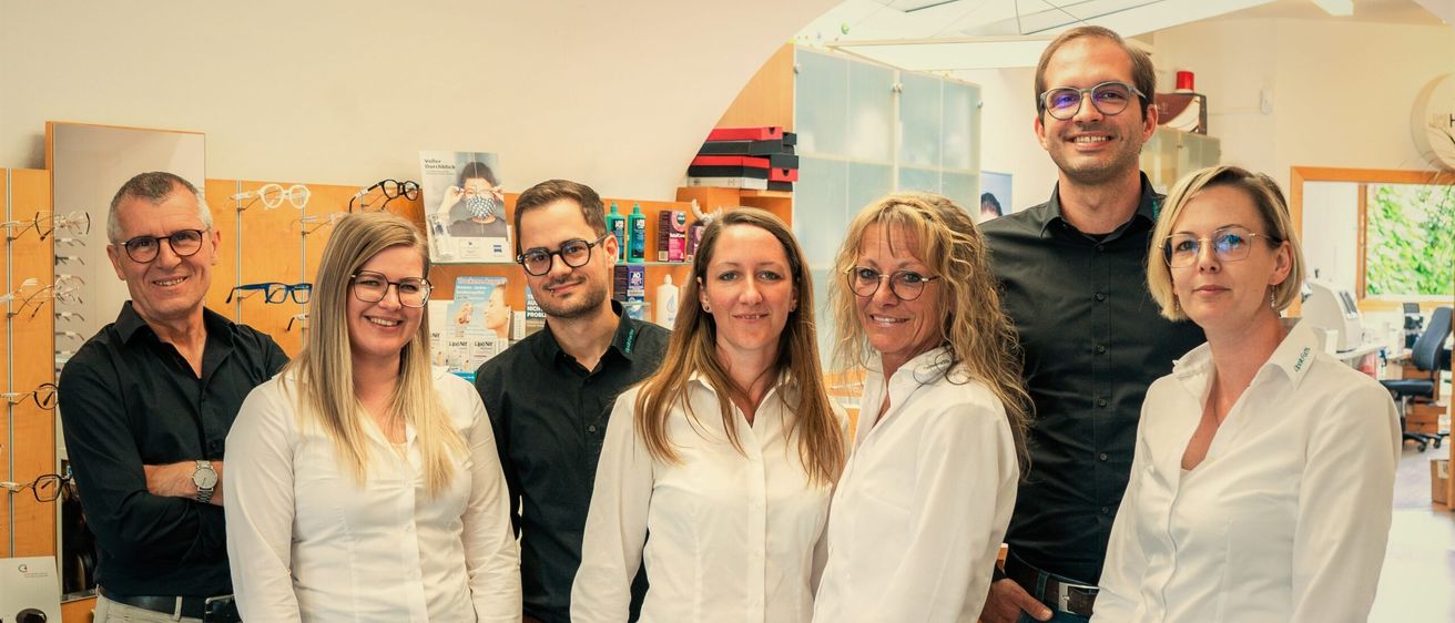 Eine Gruppe lächelnder Personen, möglicherweise Angestellte, steht zusammen in einem Büro. Sie sind in weißen Hemden und Jeans gekleidet. Hinter ihnen befinden sich Regale mit Produkten und einem Bilderrahmen.