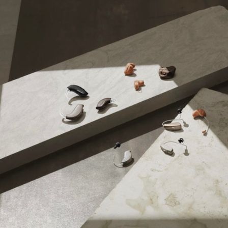 Verschiedene Modelle von Hörgeräten liegen auf einer weißen Marmoroberfläche mit einem Schatten darauf.