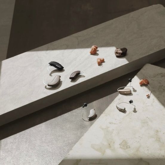 Verschiedene Modelle von Hörgeräten liegen auf einer weißen Marmoroberfläche mit einem Schatten darauf.