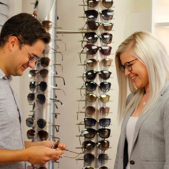 Ein Mann mit Brille hält ein Paar Sonnenbrillen und lächelt eine Frau mit blonden Haaren in einem Anzug an. Sie sind in einem Geschäft mit einer Wand voller Sonnenbrillen hinter ihnen.