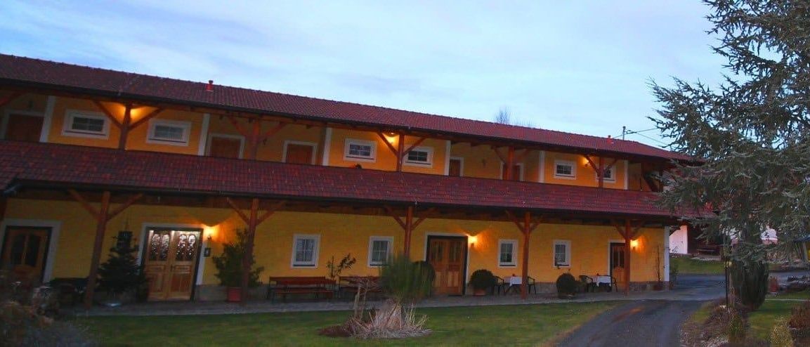 Ein großes gelbes Gebäude mit einem roten Dach und Holzsäulen. Es hat mehrere Fenster und Türen und ist von Sitzgelegenheiten im Freien und Topfpflanzen umgeben.