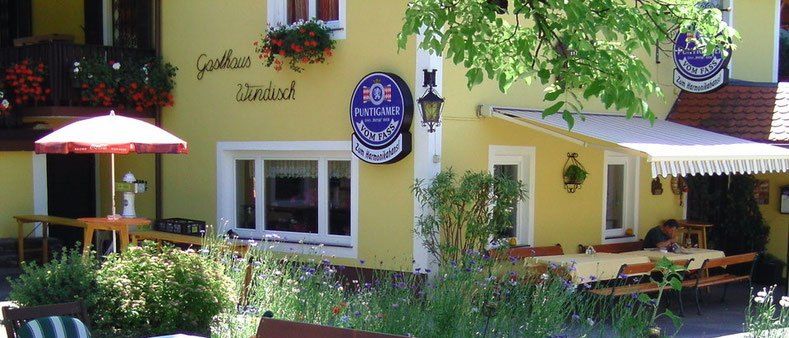 Ein gelbes Gebäude mit Sitzgelegenheiten im Freien und Tischen, die mit weißem Tuch bedeckt sind. Es gibt ein Schild mit der Aufschrift 'Puntigamer vom Fass' an der Wand.