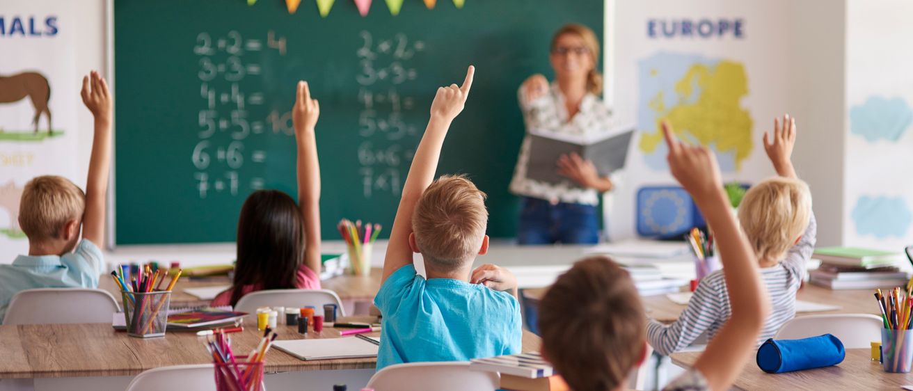 Ein Klassenzimmer mit Schülern, die Hände heben, einer Lehrerin am Tafel, und verschiedenen Schulmaterialien auf den Tischen.
