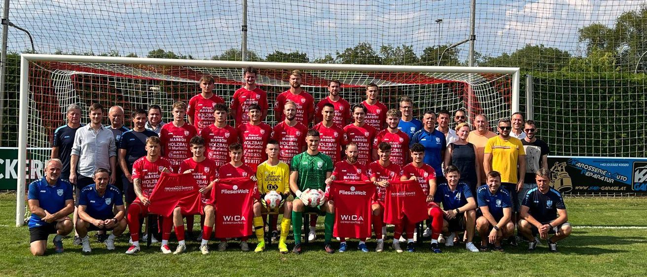 Eine Fußballmannschaft in roten Trikots posiert für ein Foto vor einem Tornetz mit roten und weißen Streifen, mit Bäumen und einem bewölkten Himmel im Hintergrund.