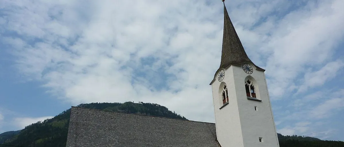 Eine weiße Kirche mit grauem Dach und einem Turm mit Uhren steht auf einem Friedhof mit Grabsteinen und einer gelben Bank. Im Hintergrund befinden sich Berge unter einem bewölkten Himmel.