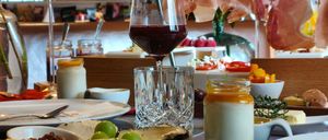 Ein Esstisch mit einer Auswahl an Speisen und Getränken, darunter Käse, Obst und Wein. Der Tisch ist mit Tellern, Gläsern und Besteck gedeckt.