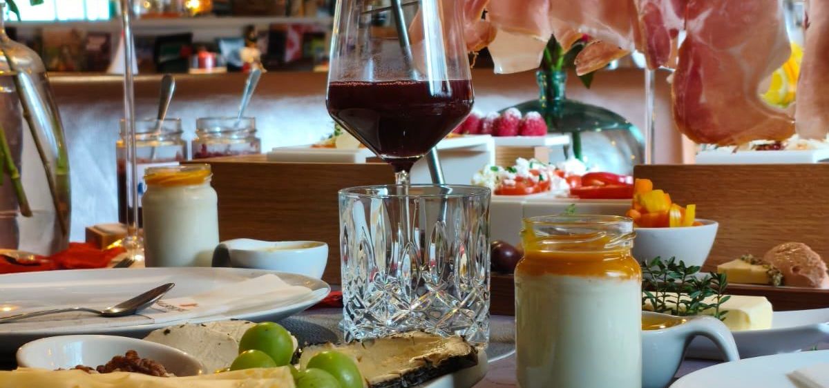 Ein Esstisch mit einer Auswahl an Speisen und Getränken, darunter Käse, Obst und Wein. Der Tisch ist mit Tellern, Gläsern und Besteck gedeckt.