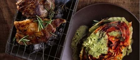 Gegrilltes Fleisch mit Rosmarin auf einem Grill, begleitet von einem Teller mit geschichtetem Gemüsegericht und einem Glas Bier auf einem Holztisch.