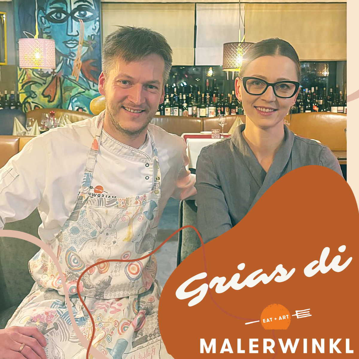 Ein Mann und eine Frau lächeln in einem Restaurant. Der Mann trägt eine Schürze und die Frau trägt eine Brille. Sie stehen nebeneinander. Dahinter befinden sich Regale mit Flaschen.