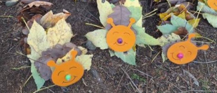 Ein Kreis aus Laubdekorationen, jede mit einem zeichentrickartigen Gesicht, aus orangefarbenem Papier gefertigt und auf dem Boden zwischen trockenen Blättern platziert.