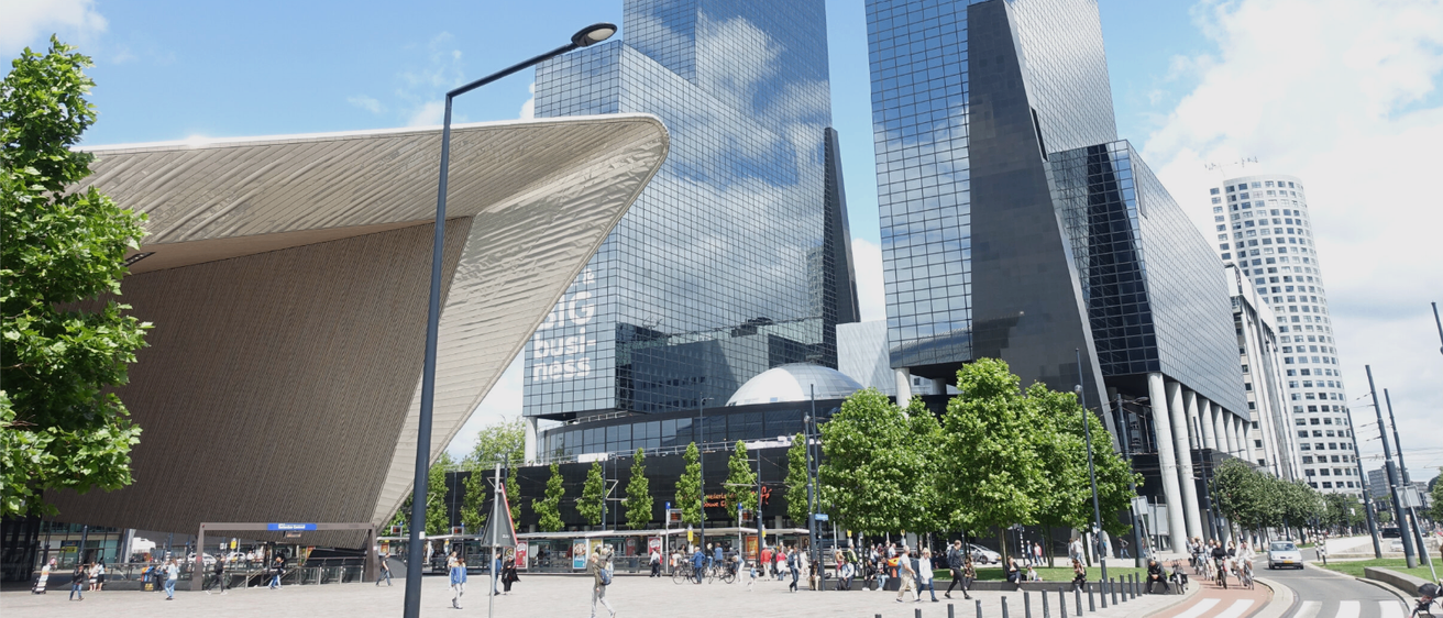 Ein geschäftiger Stadtplatz mit modernen Gebäuden, Menschen, die gehen, Straßenlaternen und Bäumen im Vordergrund. Der Himmel ist klar mit einigen Wolken.