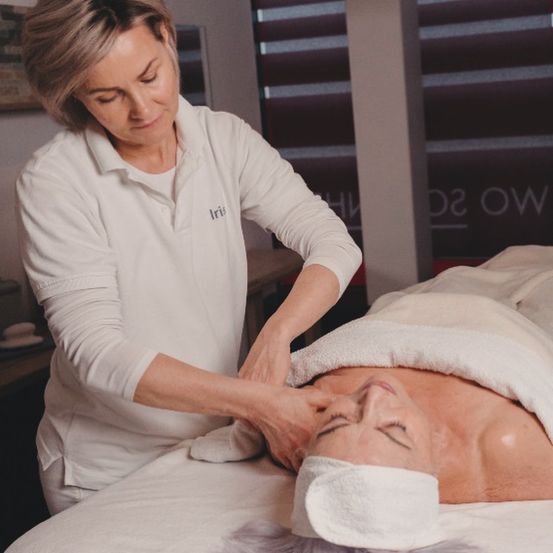 Eine Frau in einer weißen Uniform massiert den Nacken einer anderen Frau in einem Spa. Die Frau, die die Massage erhält, hat ein Handtuch um den Hals. Das Spa hat eine weiße Wand und ein schwarzes Schild.