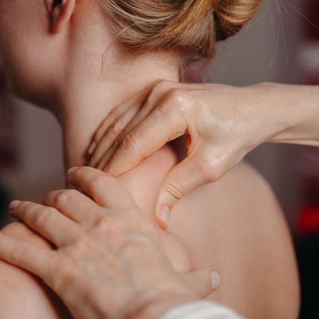 Eine Frau mit blondem Haar erhält von jemandes Händen eine Nackenmassage. Die Hände der Person liegen auf dem Nacken der Frau.