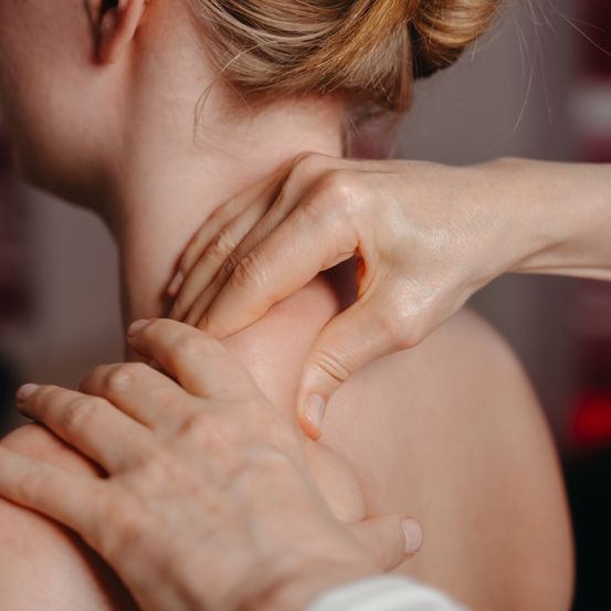 Eine Frau mit blondem Haar erhält von jemandes Händen eine Nackenmassage. Die Hände der Person liegen auf dem Nacken der Frau.
