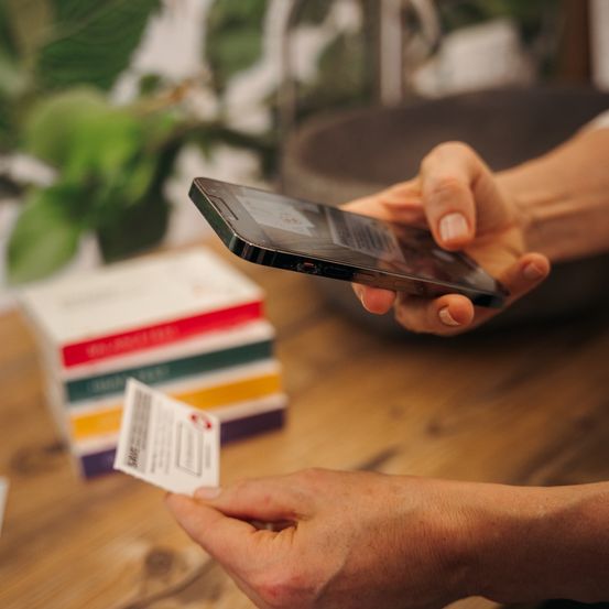 Eine Person hält ein Smartphone und eine Karte und scannt möglicherweise einen QR-Code auf der Karte. Die Karte liegt auf einem Holztisch mit bunten Schachteln dahinter.