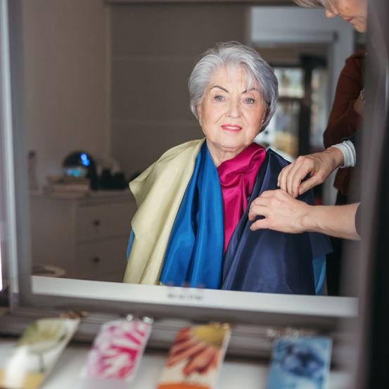 Eine ältere Frau mit weißen Haaren betrachtet sich im Spiegel. Sie trägt einen mehrfarbigen Schal. Eine andere Person justiert ihren Schal. Im Spiegel sehen wir eine Kommode, eine Lampe und eine Tür.