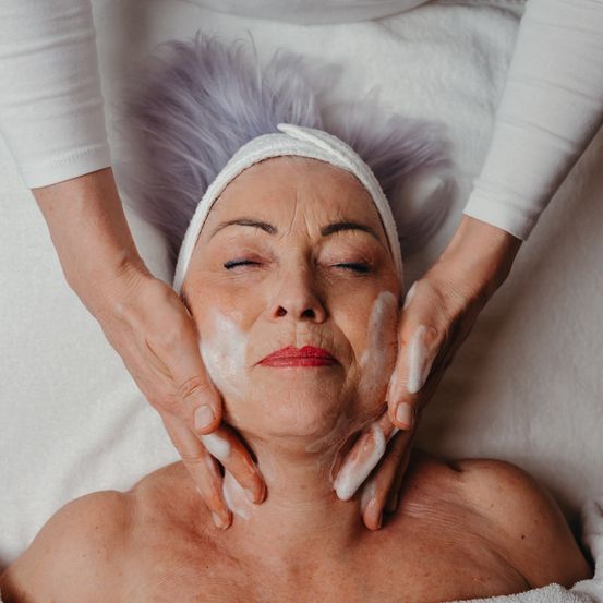 Eine ältere Frau liegt auf einem weißen Handtuch und bekommt von einer Person mit lila Haaren eine Gesichtsbehandlung. Das Gesicht der Frau ist mit weißer Creme bedeckt und ihre Augen sind geschlossen.
