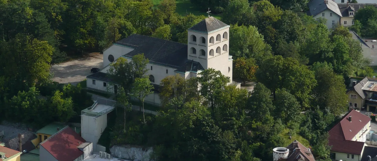 Ein Luftbild einer Kirche, umgeben von üppigem Grün und Bäumen, mit einem Turm und einem Kreuz auf der Spitze sichtbar.