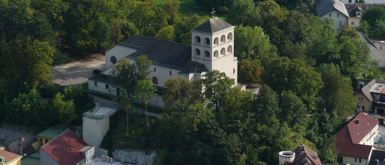 Ein Luftbild einer Kirche, umgeben von üppigem Grün und Bäumen, mit einem Turm und einem Kreuz auf der Spitze sichtbar.