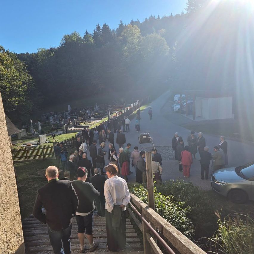 Trauergemeinde versammelt sich am Friedhof in einer ländlichen Umgebung bei Sonnenschein