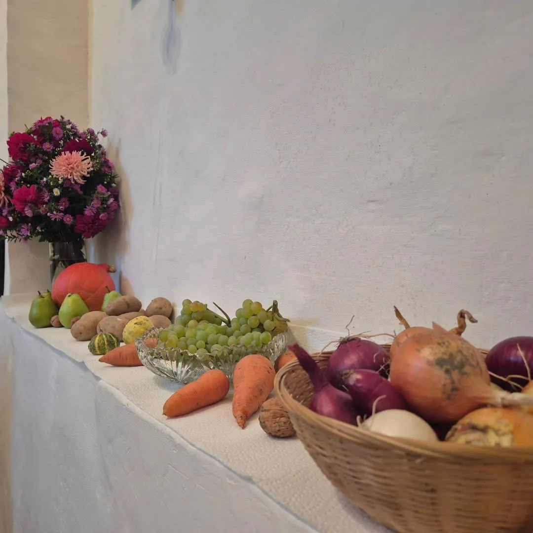 Eine weiße Wand mit einer Vielzahl von Obst und Gemüse zur Schau gestellt. Links steht ein Blumenstrauß und rechts ein Korb mit Zwiebeln.