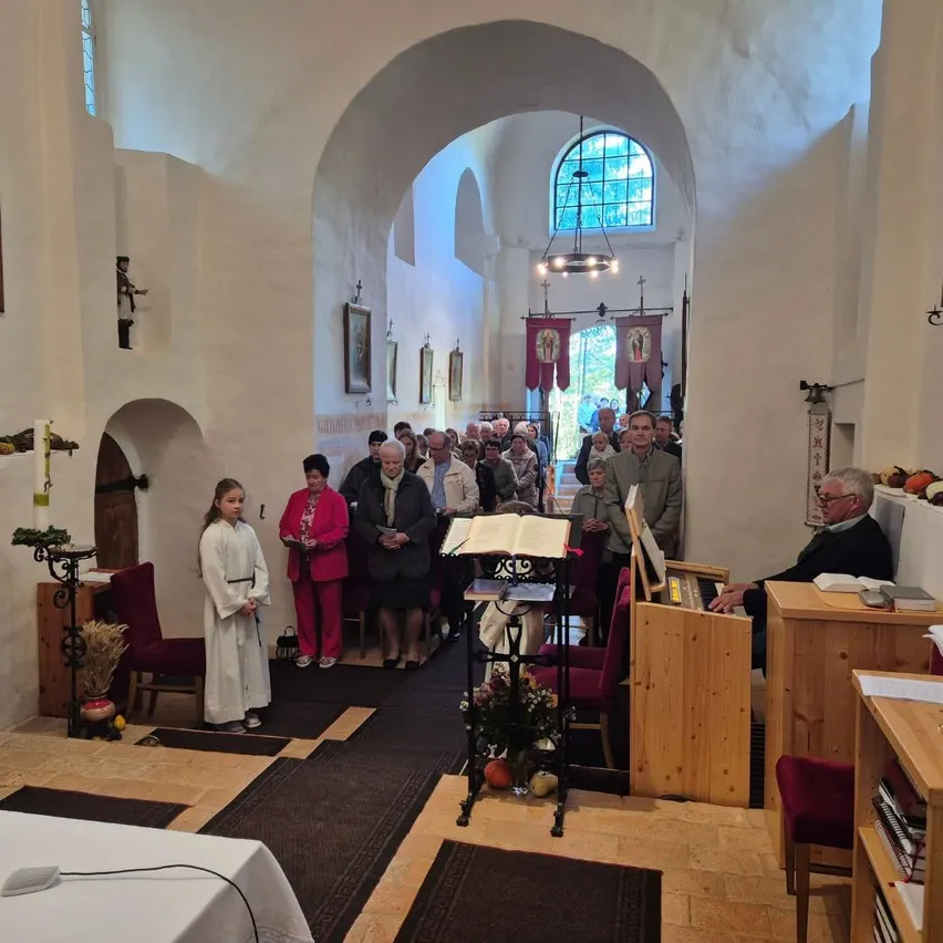 Eine Gruppe von Menschen ist in einer Kirche versammelt, möglicherweise für eine religiöse Zeremonie. Ein junges Mädchen in einem weißen Gewand steht vorne, und ein älterer Mann spielt die Orgel.