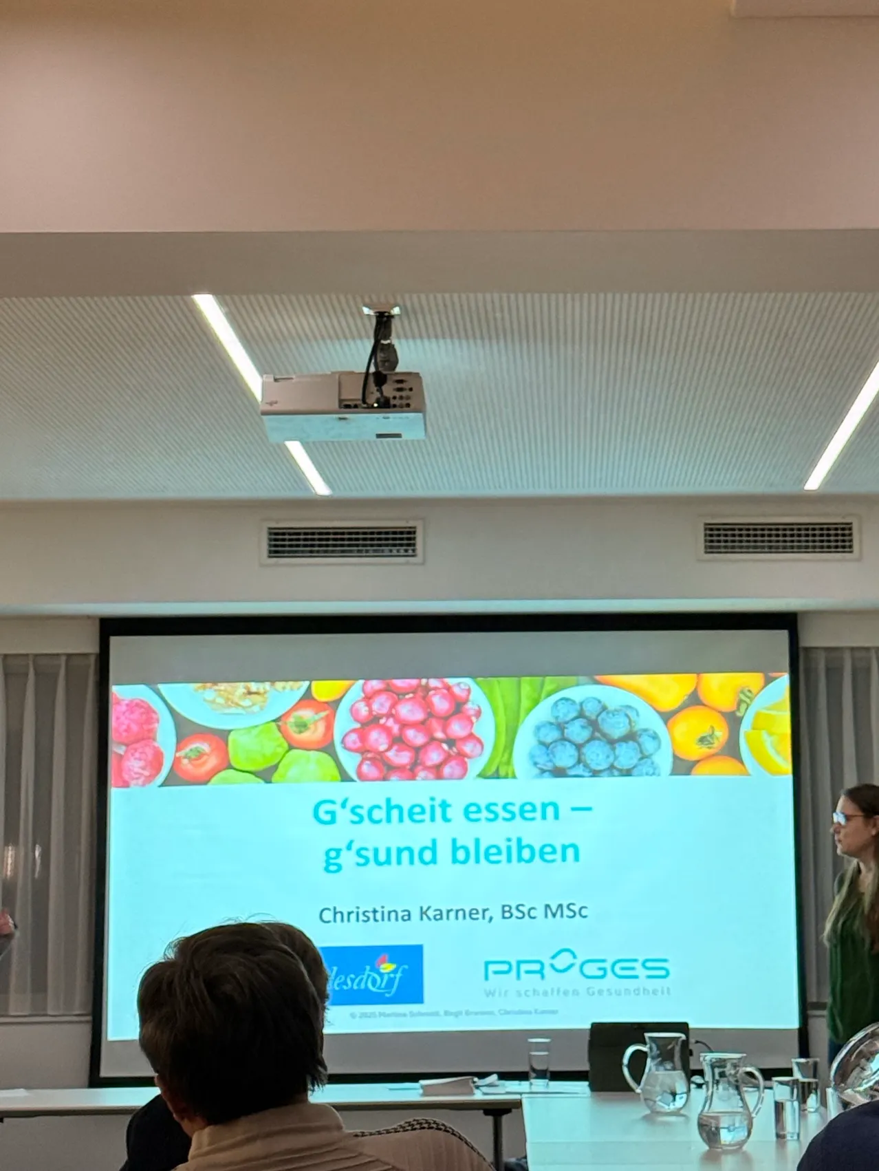 Eine Person sitzt vor einer Projektionsleinwand in einem Raum. Die Leinwand zeigt eine Präsentation mit dem Titel 'G'sheit essen - g'sund bleiben' von Christina Karner. Die Präsentation enthält Bilder von Obst und Gemüse. Eine Frau steht neben der Leinwand.