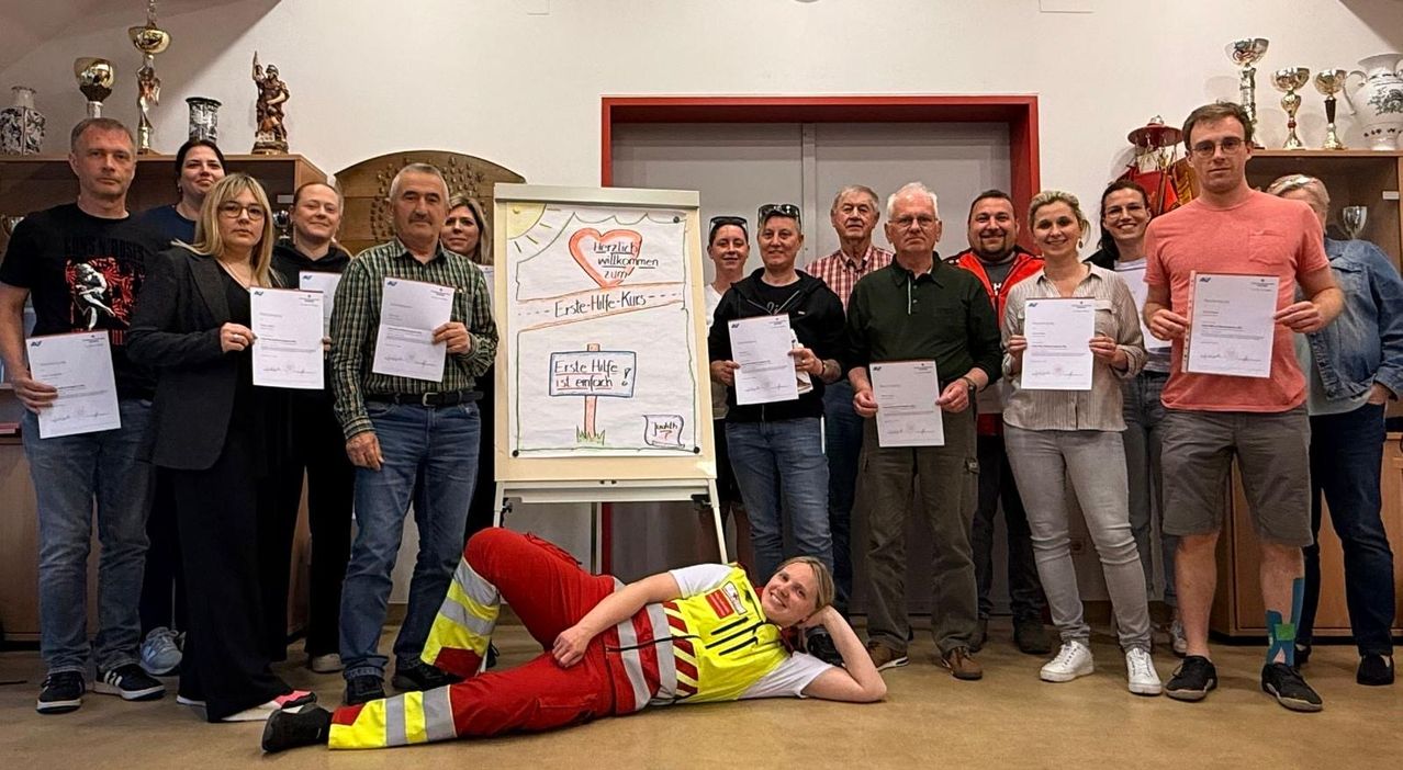 A group of people stand behind a board with a heart and text, holding papers. One person lies on the ground with a smile.