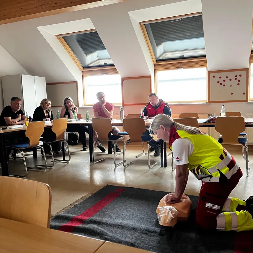 Eine Gruppe von Menschen sitzt an einem Tisch in einem Klassenzimmer. Eine Frau in einer gelben Weste kniet auf einem Teppich und demonstriert Herz-Lungen-Wiederbelebung an einem Puppenkörper.