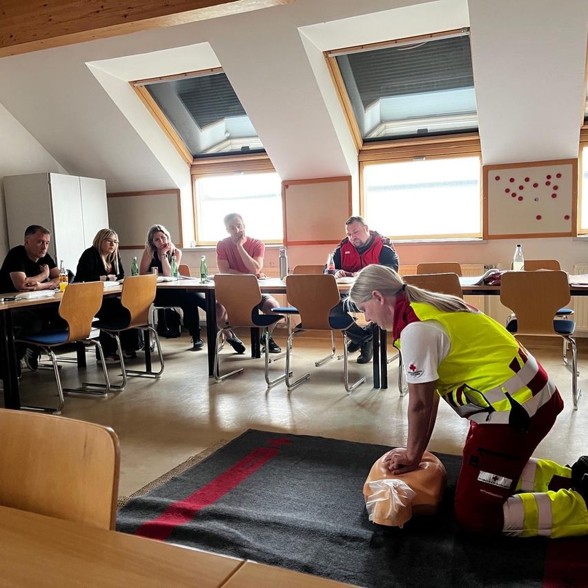 A group of people are seated at a table in a classroom setting. A woman in a yellow vest is kneeling on a mat and demonstrating CPR on a dummy.