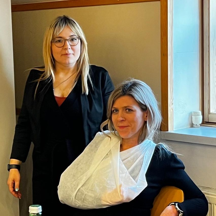 Two women stand in an indoor setting. One with a bandaged arm, the other with glasses and a necklace. Behind them, a window with a white object on the sill.
