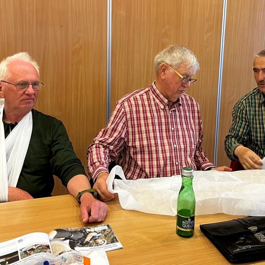 Three men sit around a table. One man has a bandaged arm. Another man holds a plastic bag. There is an open book and a bottle on the table.