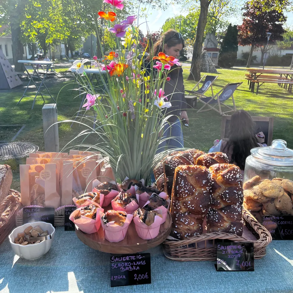 Ein Tisch ist mit Gebäck, Brot und Keksen gefüllt. Eine Blumenarrangement steht in der Mitte. Eine Frau steht hinter dem Tisch. Es gibt andere Tische, Bänke und Bäume im Hintergrund.