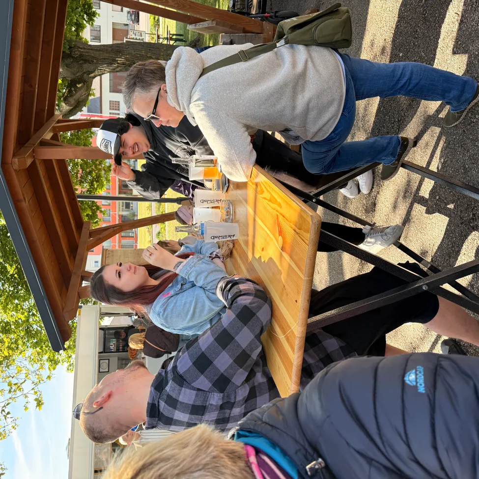 Eine Gruppe von Menschen versammelt sich um einen Tisch im Freien und genießt Getränke. Eine Person sitzt, während andere stehen. Bäume und Gebäude sind im Hintergrund.