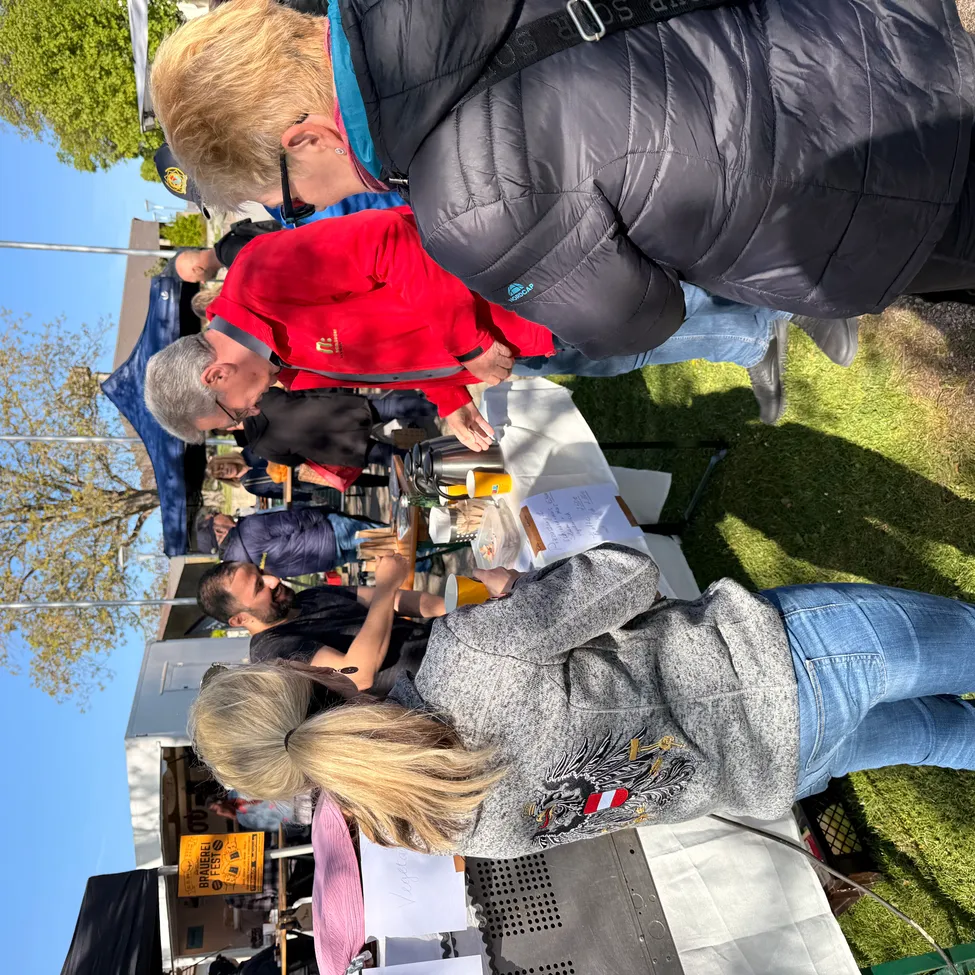 Eine Gruppe von Menschen versammelt sich an einem Tisch mit Essen und Getränken an einem sonnigen Tag. Einige tragen Jacken und Brillen. Eine Frau mit blondem Haar steht vorne und schaut einen Mann an. Ein Schild ist in der Nähe, und es gibt Zelte und Bäume in der Ferne.