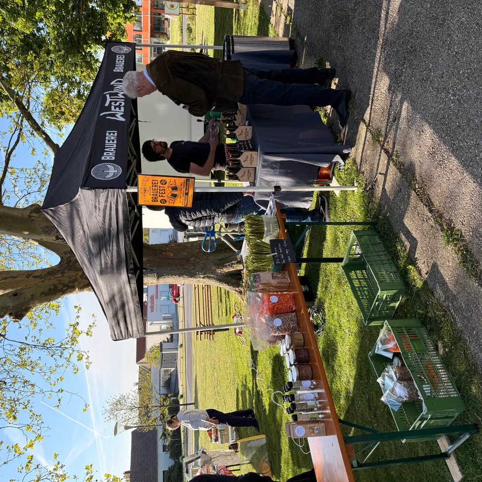 Ein Outdoor-Markt mit einem Zelt und verschiedenen Artikeln auf Tischen. Menschen stehen und einer sitzt. Grüne Kisten mit Produkten. Ein Schild mit 'Brauere Westland'.